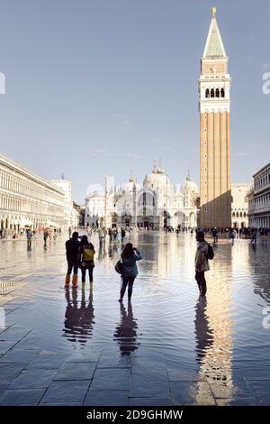 Touristen fotografieren auf dem Markusplatz bei Flut, Acqua alta. Der Markusdom und der Glockenturm spiegeln sich auf dem Wasser. Stockfoto