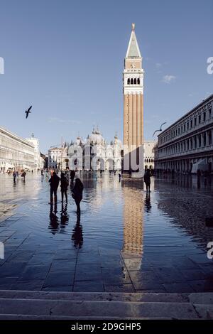 Touristen fotografieren auf dem Markusplatz bei Flut, Acqua alta. Der Markusdom und der Glockenturm spiegeln sich auf dem Wasser. Stockfoto