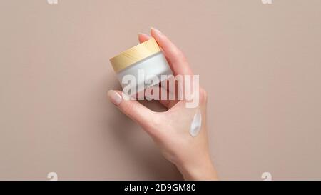 Weibliche Hand halten Glas kosmetische Creme auf pastellbeige Hintergrund. Frau Anwendung Feuchtigkeitscreme Stockfoto