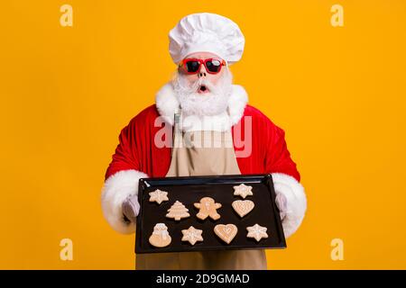 Foto von santa claus aufgeregt Großvater grauen Bart halten bereit Ingwer Brot Süßigkeiten Haushalt Aufgaben Kochen süchtig tragen Kostüm Handschuhe Sun Specs Kappe Stockfoto
