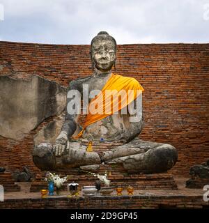 Eine sitzende buddha-Statue, die eine Safran-Schärpe in einem Backsteinhof eines thailändischen buddhistischen Tempels trägt. Vor der Statue gibt es eine Vielzahl von Opfergaben Stockfoto