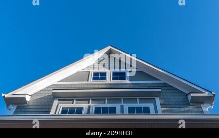 Dekorativer Giebel mit weißen Dachrinnen und Soffit, grauen Schindelschiefern, Ausstoßfenstern, eingerahmt von einem dicken weißen Rahmen mit blauem Himmel auf einer neuen Konstruktion Stockfoto