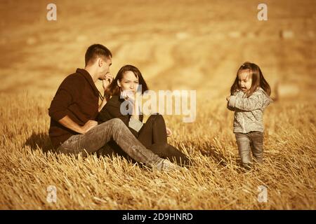 Glückliche junge Familie mit zwei Jahre alten Baby Mädchen sitzen Auf dem Boden in geerntetem Feld Stockfoto