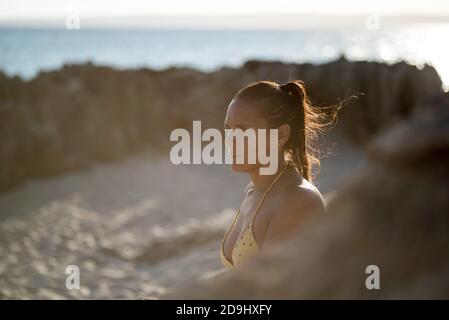 Nahaufnahme einer hübschen Frau gegen das Licht im Bikini, die an einem Strand mit dem Meer im Hintergrund zur Seite schaut, nachdenklich und melancholisch aussieht. Stockfoto