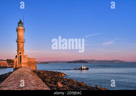 Fischerboot vorbei am Leuchtturm, verlassen alten venezianischen Hafen von Chania, Kreta, Griechenland bei Sonnenaufgang. Am frühen Morgen Himmel und kretische Hügel in der Ferne. Stockfoto