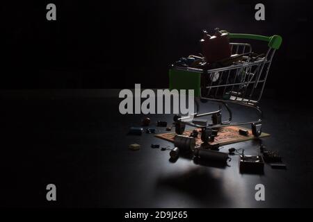 Metallwagen Warenkorb, gefüllt mit Funkkomponenten, steht auf einer elektronischen Platine. Foto in dunkler Hintergrundbeleuchtung. Stockfoto