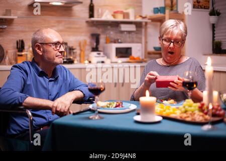 Alte Frau schaut überrascht halten Geschenk von Ehemann im Rollstuhl während des festlichen Abendessens. Fröhliche fröhliche ältere Paar zusammen zu Hause essen, genießen Sie das Essen, feiern ihre Ehe, Stockfoto
