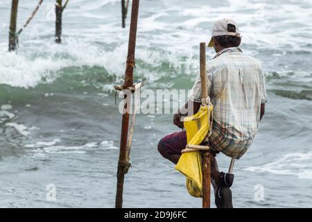 Stelzenfischer mit seiner Holzstange, die mit seiner gelben Tasche an der Stange zurück zur Kamera zeigt, fischt in Sri Lanka in einer traditionellen, einzigartigen Methode Stockfoto