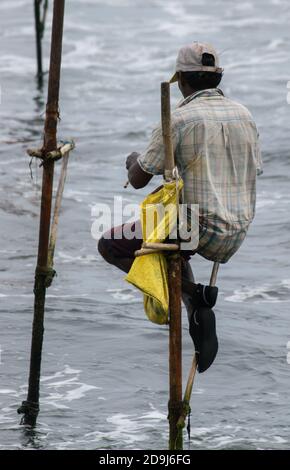 Stelzenfischer mit seiner Holzstange, die mit seiner gelben Tasche an der Stange zurück zur Kamera zeigt, fischt in Sri Lanka in einer traditionellen, einzigartigen Methode Stockfoto