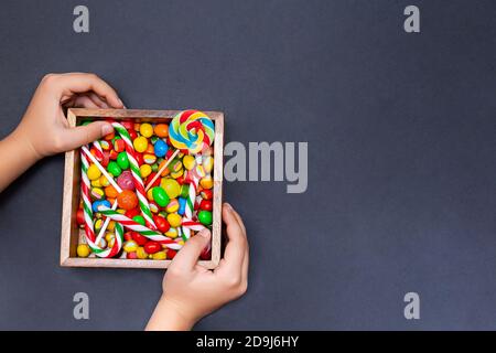 Eine Schachtel Süßigkeiten in den Händen eines Jungen auf schwarzem Hintergrund. Verschiedene farbige Bonbons, Lutscher und Marmelade in einer Holzkiste, Draufsicht, Kopierraum. Stockfoto