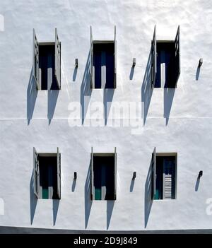 Offene, graue Holzläden in einem mediterranen Gebäude aus weißem Stein mit scharfen Schatten, die von der hohen Mittagssonne erzeugt werden. Stockfoto
