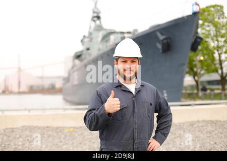 Porträt des Kapitäns der Marine, der in der Nähe eines großen Schiffes im Hintergrund steht und Daumen nach oben zeigt. Stockfoto