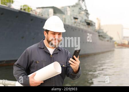 Marine lächelnde Ingenieur im Gespräch mit VHF Walkie Talkie, hält Papiere in der Nähe des Schiffes im Hintergrund. Stockfoto