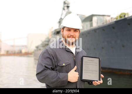 Kapitän liest Vertrag auf Tablet und zeigt Daumen nach oben in der Nähe von Schiff im Hintergrund. Stockfoto