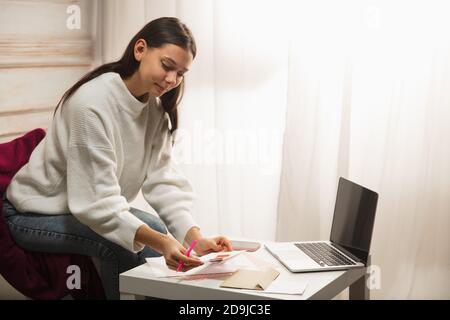 Komfortabel. Frau Schreiben Brief, Grußkarte für Neujahr und Weihnachten 2021 für Freunde oder Familie sitzen zu Hause mit Laptop. Feiertage, Feier. Sieht fröhlich, glücklich, vorsichtig aus. Stockfoto