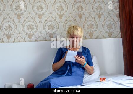 Entspannte Frau sitzt auf dem Bett im Hotel, hält ein Smartphone oder online einkaufen, um mobile Anwendung oder soziales Netzwerk zu überprüfen. Stockfoto