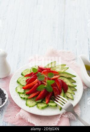 Veganer Salat aus Erdbeere, Gurke und Avocado mit Mohn, Öldressing und Minze. Frische, gesunde, diätetische Lebensmittel. Auf einem weißen Teller und auf einer blu Stockfoto