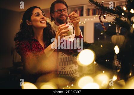 Paar Dekoration Weihnachtsbaum bei Familie zu Hause. Frau binden glänzenden dekorativen Ball auf Weihnachtsbaum mit Mann stehen. Stockfoto
