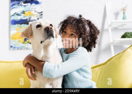 Lächelndes afroamerikanisches Mädchen, das labrador umarmt und anschaut, während es zu Hause auf dem gelben Sofa sitzt, auf verschwommenem Hintergrund Stockfoto