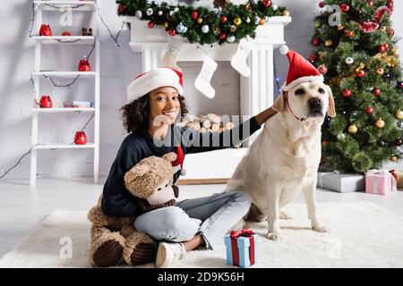 Lächelndes afroamerikanisches Mädchen streichelte labrador Hund, während er auf saß Boden mit Teddybär Stockfoto