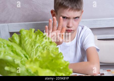 Ein kaukasischer Junge weigert sich, Salat zu essen, macht mit offener Hand ein Stoppschild mit einem ernsten und selbstbewussten Ausdruck auf seinem Gesicht, eine Geste des Schutzes. Das Konzept der nützlichen und ungesunden Lebensmittel Stockfoto
