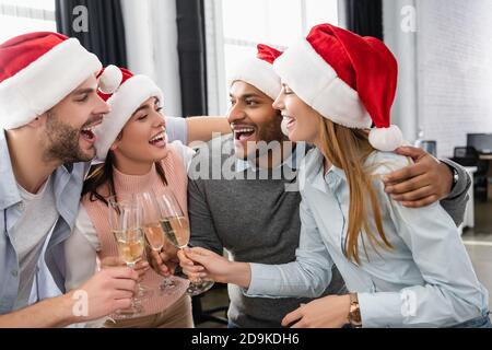 Multiethnische Geschäftsleute in Santa-Hüten umarmen beim Klatschen mit Champagner Im Büro Stockfoto