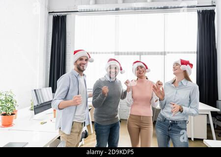 Fröhliche multiethnische Geschäftsleute in santa Hüte zeigen yeah Geste in Büro Stockfoto