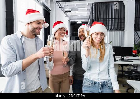 Positive multiethnische Geschäftsleute in weihnachtsmützen mit Gläsern Champagner Im Büro Stockfoto