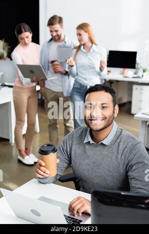Lächelnder indischer Geschäftsmann hält Kaffee, um zu gehen, während er Laptop benutzt In der Nähe von Kollegen auf unscharfem Hintergrund Stockfoto