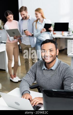 Indischer Geschäftsmann lächelt an der Kamera, während er einen Laptop in der Nähe von Kollegen benutzt Auf unscharfem Hintergrund im Büro Stockfoto