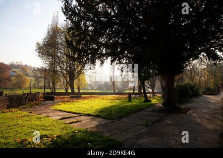 Sonnenlicht durchdringt die bunten Herbstbäume in und um einen abgelegenen Kirchhof in Kirkheaton Village in West Yorkshire, England Stockfoto