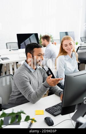 Indischer Geschäftsmann zeigt auf Computer in der Nähe lächelnden Kollegen im Büro Stockfoto