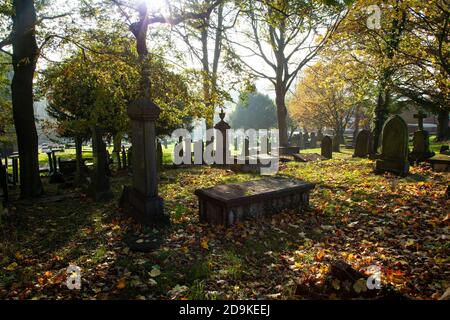 Ein Yorkshire Friedhof mit Sonne, die durch die Baumkronen bricht und das gefallene Herbstlaub mit getupftem Licht beleuchtet, in der Nähe von Huddersfield, Großbritannien Stockfoto