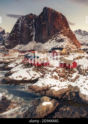 Ikonische rote Stelzenhäuser an der Küste im Dorf Hamnøy auf den Lofoten-Inseln in Norwegen bei klarem Winter Morgen mit schneebedeckten steilen Berg Stockfoto