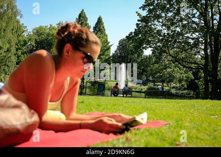 Essen, Nordrhein-Westfalen, Ruhrgebiet, Deutschland, Essener Stadtgarten, junge Frau mit Sonnenbrille liegt auf der Wiese und liest ein Buch, Parklandschaft mit See und Wasserbrunnen anlässlich der Essen 2017 grünen Hauptstadt Europas. Stockfoto