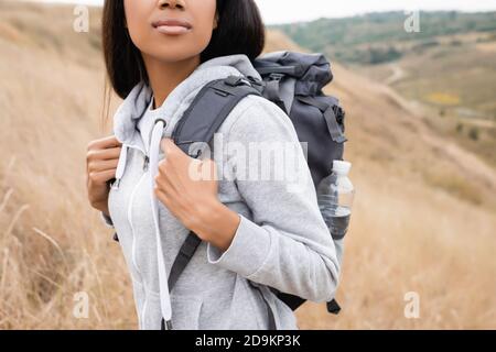 Beschnittene Ansicht der afroamerikanischen Frau mit Rucksack stehend auf Hügel mit verschwommener Landschaft im Hintergrund Stockfoto