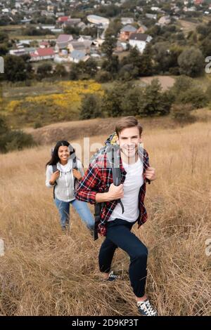 Junger Tourist mit Rucksack lächelnd an der Kamera in der Nähe von afroamerikaner Frau auf verschwommenem Hintergrund im Freien Stockfoto