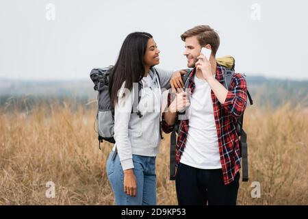 afroamerikanische Frau in der Nähe von Freund im Gespräch auf Smartphone und Berühren Rucksack auf Rasen Stockfoto