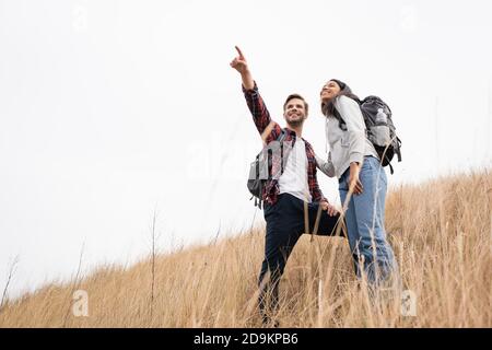 Low-Winkel-Ansicht des lächelnden Mannes mit Rucksack zeigt mit Finger weg in der Nähe afroamerikanische Frau, während sie auf Gras steht Hügel mit Himmel im Hintergrund Stockfoto