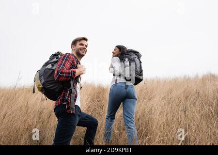 Junge Wanderer lächelnd in der Nähe afroamerikanische Freundin zu Fuß auf Gras Hügel Stockfoto
