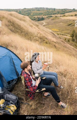 Hochwinkel Blick auf lächelnde multiethnische Touristen mit Tasse und Thermoskannen in der Nähe von Rucksäcken und Zelt auf Rasen Stockfoto