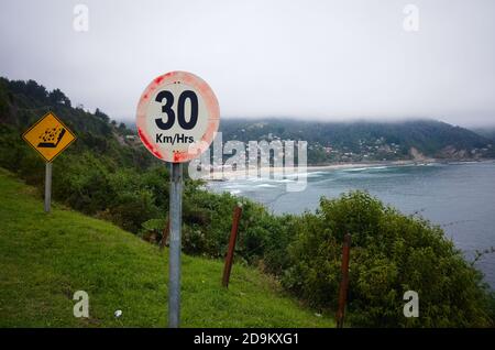 Straßenschild Geschwindigkeitsbegrenzung auf einer Straße in den Bergen mit scharfer Kurve. Steine fallen Warnung Verkehrsschild. Meer Strand und Dorf im Hintergrund. Chile Stockfoto