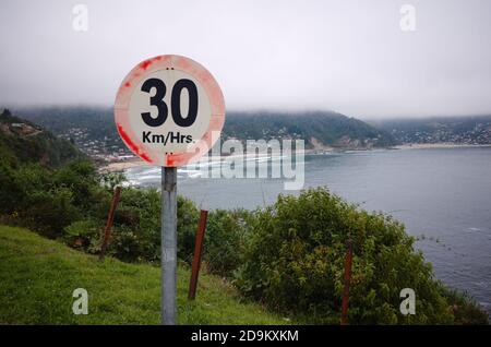 Geschwindigkeitsbegrenzungszeichen auf einer scharfen Kurve vor Klippe. 30 km pro Stunde Geschwindigkeitsbegrenzung auf Bergstraße. Meeresstrand und kleines Dorf im Nebel im Hintergrund. Stockfoto