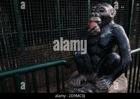 Lustige Statue des schwarzen Schimpansen in der Nähe von Affenkäfigen in einem Zoo. Steinaffe sitzt auf einem Stumpf essen roten Apfel und begrüßt Touristen und Besucher Stockfoto