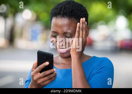 Junges Mädchen, das in blauer Kamisole steht und auf das Mobiltelefon schaut, das mit der Hand auf der Wange lacht. Stockfoto