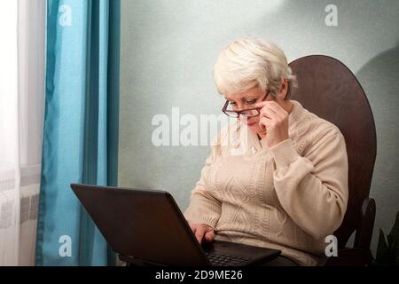 Eine ältere Dame sitzt zu Hause in einem Stuhl mit einem Laptop, hält ihre Brille und Drucke auf der Tastatur Stockfoto