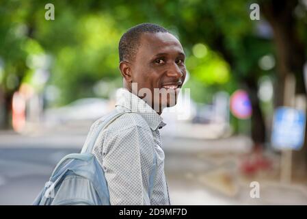 Junger Mann steht mit Rucksack und schaut lächelnd auf die Kamera. Stockfoto