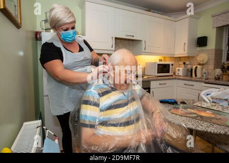 Älterer Mann in den Achtzigern, der sich von einem Friseur mit PSA als Schutz vor Coronaviren bei der Arbeit aus der Ferne schneiden lässt, England, Großbritannien Stockfoto