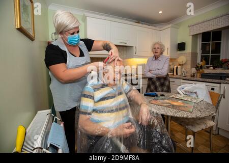 Älterer Mann in den Achtzigern, der sich von einem Friseur mit PSA als Schutz vor Coronaviren bei der Arbeit aus der Ferne schneiden lässt, England, Großbritannien Stockfoto