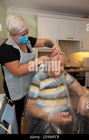 Älterer Mann in den Achtzigern, der sich von einem Friseur mit PSA als Schutz vor Coronaviren bei der Arbeit aus der Ferne schneiden lässt, England, Großbritannien Stockfoto